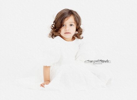 little girl in white dress looking at camera sat on the floor of white background legs out in front