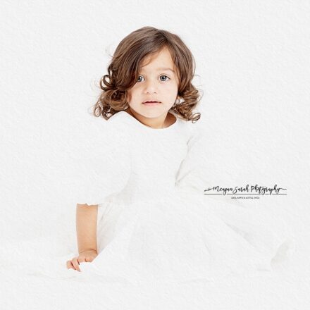 little girl in white dress looking at camera sat on the floor of white background legs out in front