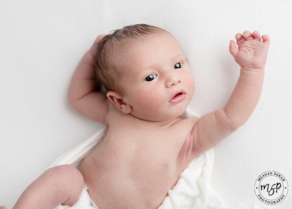 newborn baby boy laid on white sheet with arms up over head looking off camera slightly