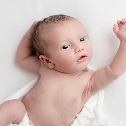 newborn baby boy laid on white sheet with arms up over head looking off camera slightly