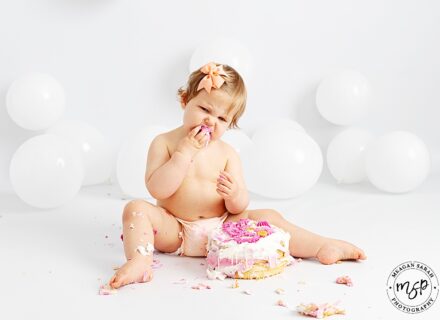 little girl on white background with peach bow at side of her hair holding cake and eating it