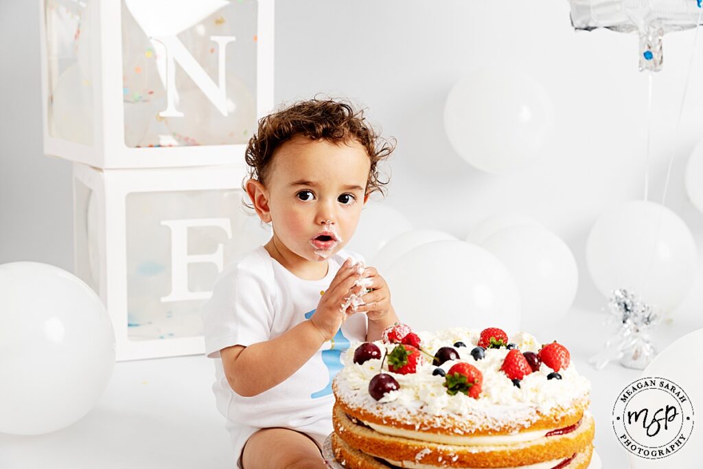 little boy on white background with white balloons and boxes spelling out one, with a Victoria sponge cake with strawberries. he is eating the strawberries legs either side of cake look to camera
