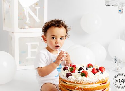 little boy on white background with white balloons and boxes spelling out one, with a Victoria sponge cake with strawberries. he is eating the strawberries legs either side of cake look to camera