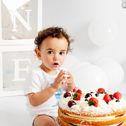 little boy on white background with white balloons and boxes spelling out one, with a Victoria sponge cake with strawberries. he is eating the strawberries legs either side of cake look to camera