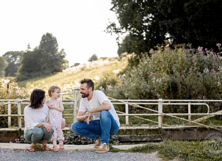 mum dad and little girl crunched down outside with a field behind them with lots of trees