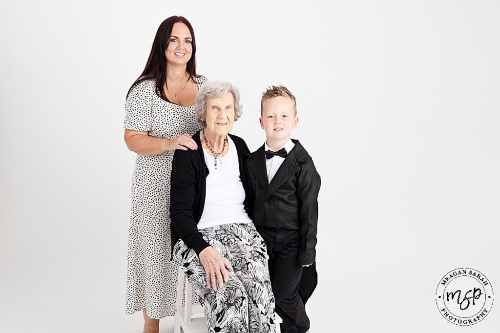 mum, little boy and great grandma against a white background smiling and facing the camera. great grandma sat down on stool in front of mum with son to the side of them