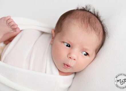 close up of newborn baby girls face swaddled I white sheet and she is pouting lookingoff camera