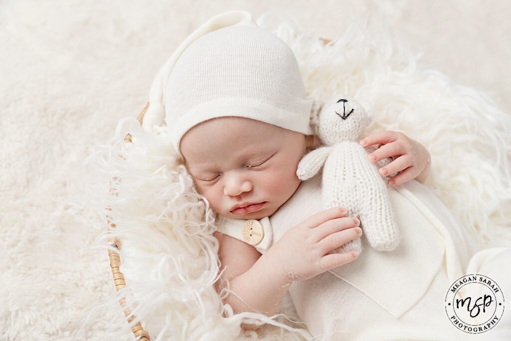 newborn baby boy in a basket dressed in cream with cream fur throw in basket baby boy holding cream teddy bear with a cream hat on asleep