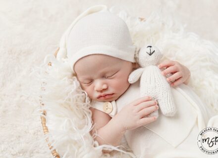 newborn baby boy in a basket dressed in cream with cream fur throw in basket baby boy holding cream teddy bear with a cream hat on asleep