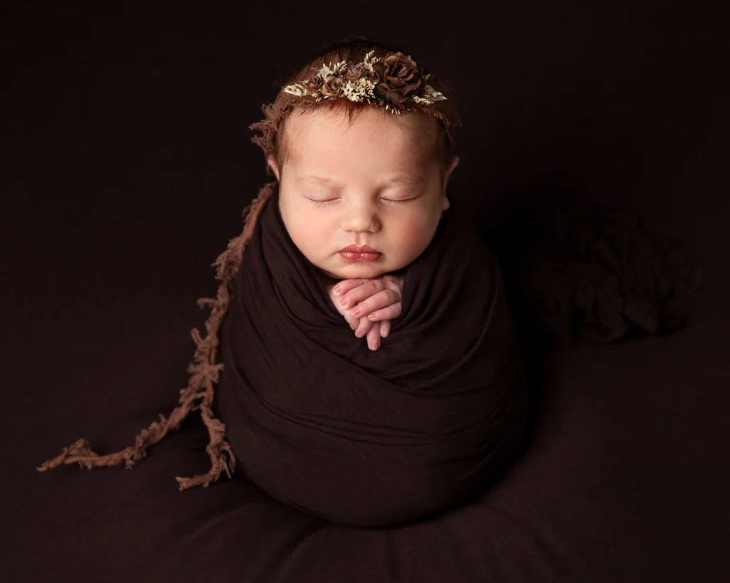 newborn baby asleep on brown background all swaddled up.