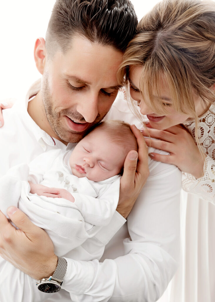 Baby photography Leeds. Baby photographer. Newborn photograph of family of three in our Leeds studio. White background, dressed in white, looking at their baby. High key.