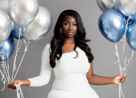 A beautiful woman with long dark hair and a white dress holding blue and white balloons. A birthday shoot. Grey backdrop. Photographed in a professional studio.