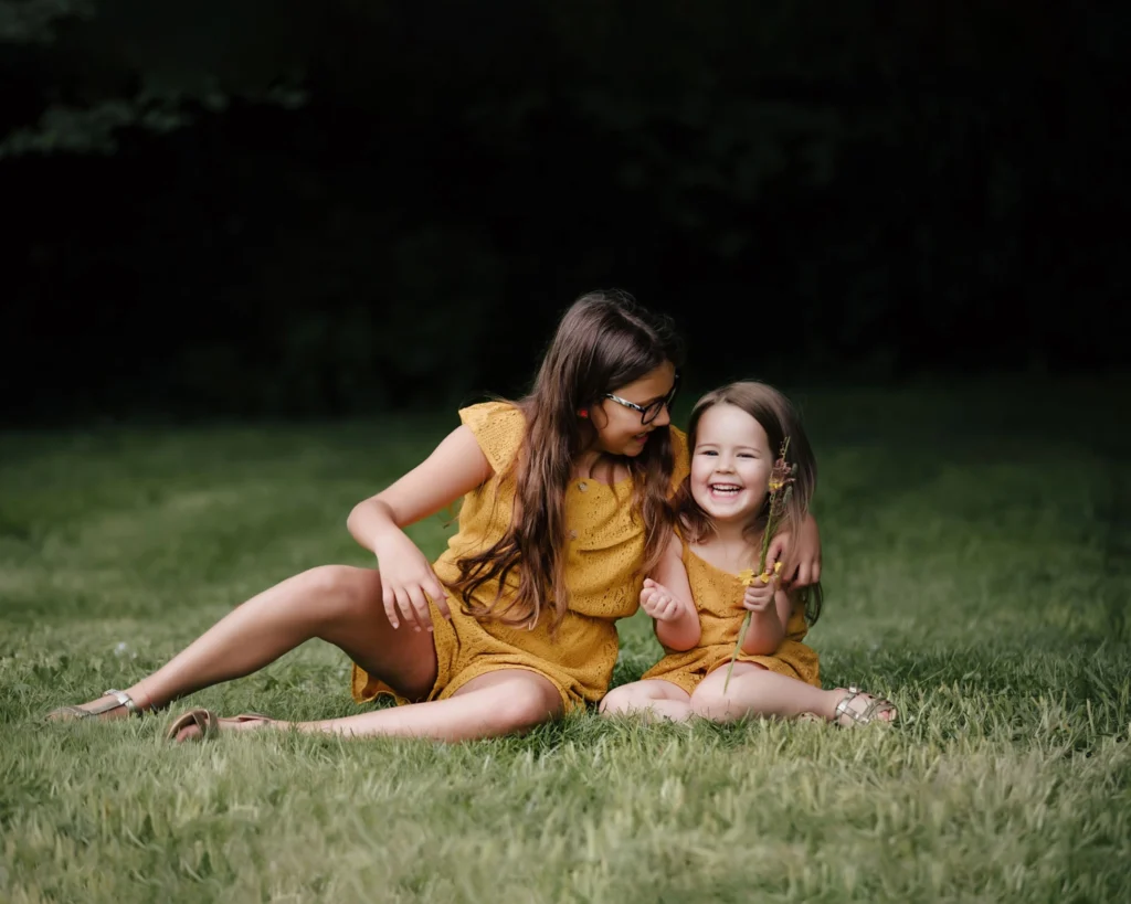 summer garden photoshoot. two sisters sitting hugging in yellow holding flowers.