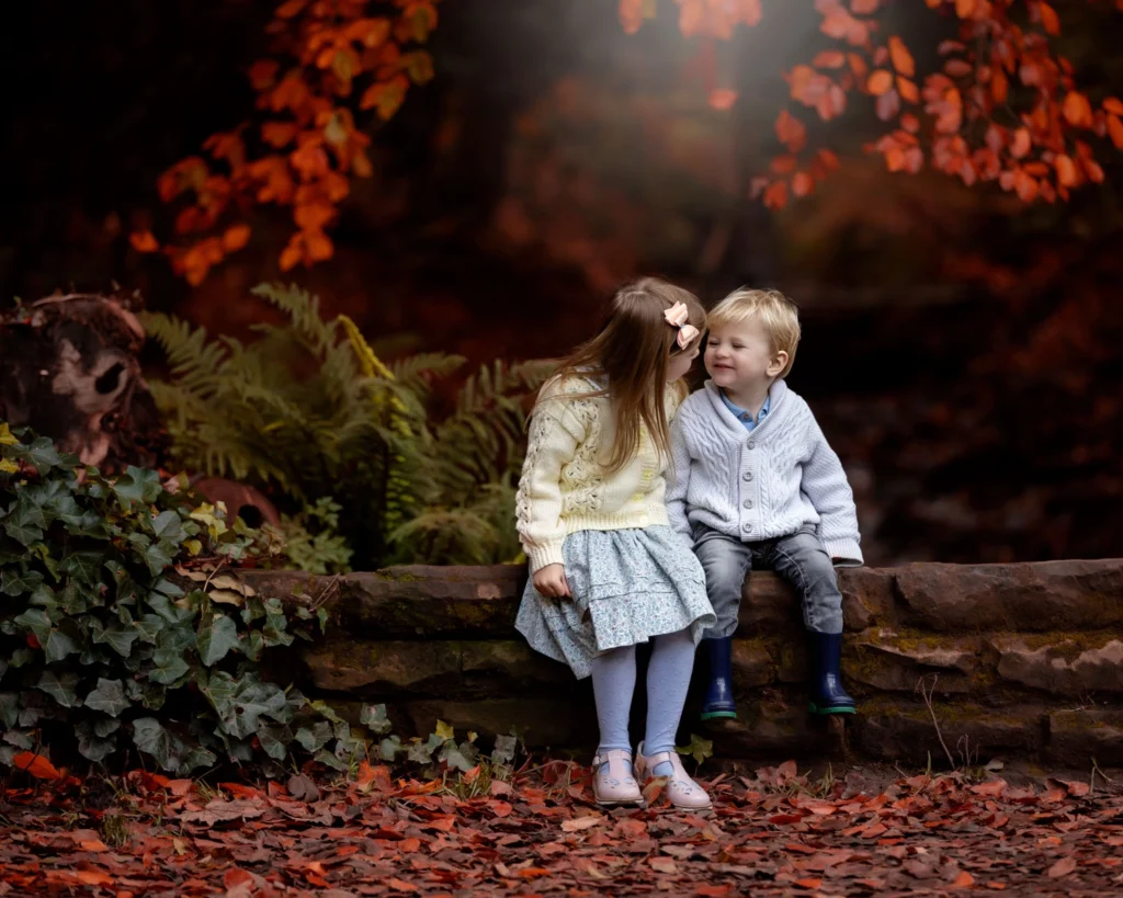 autumn photoshoot at roundly park Leeds. Boy and. girl looking at each other sitting on a wall surrounded by rich red autumn leaves.