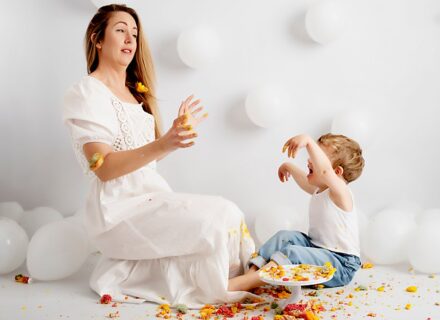 Funny cake smash photography. Mother and son smashing a cake on a white set with white balloons. Colourful rainbow cake. Laughing and being silly.