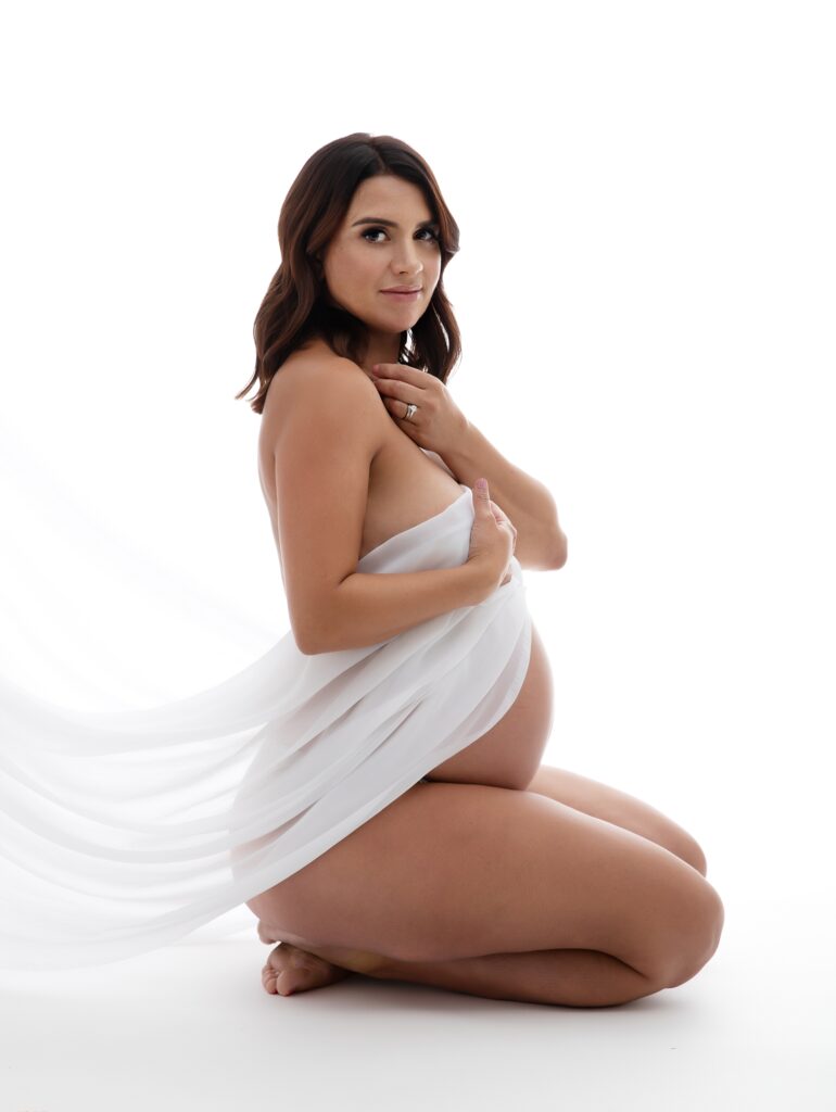 maternity photography. Pregnant Leeds photoshoot. Beautiful brunete woman with white fabric draped over her pregnancy bump. All white studio. Light and airy feel.