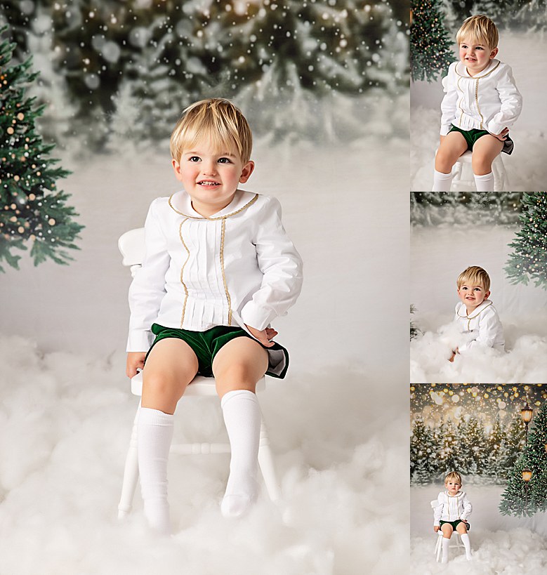 Little blonde smiling boy dressed in white shirt with gold detail and velvet blue shorts. Sitting on a small white chair surrounded by faux white snow. Background is snowy christmas tree scene. Photographed in a photography studio.