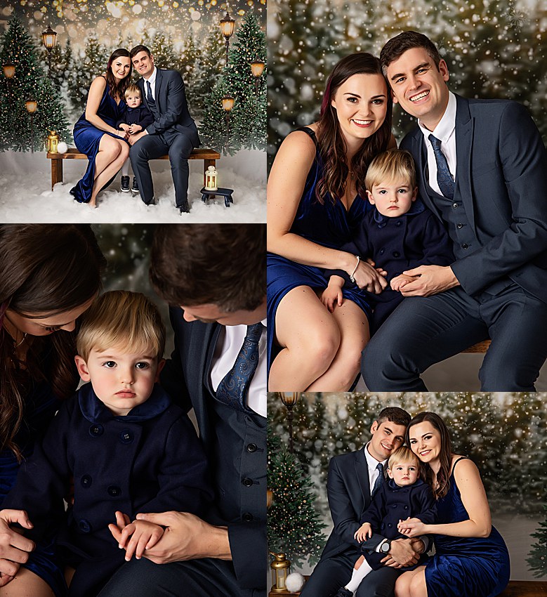 Christmas photography set. Family of 3 posing against a dark, street lamp lit woodlands covered in snow. People wearing all dark royal, navy blue. Dressed smartly. Smiling at the camera. Sitting on a wooden bench. Surrounded by faux white snow.
