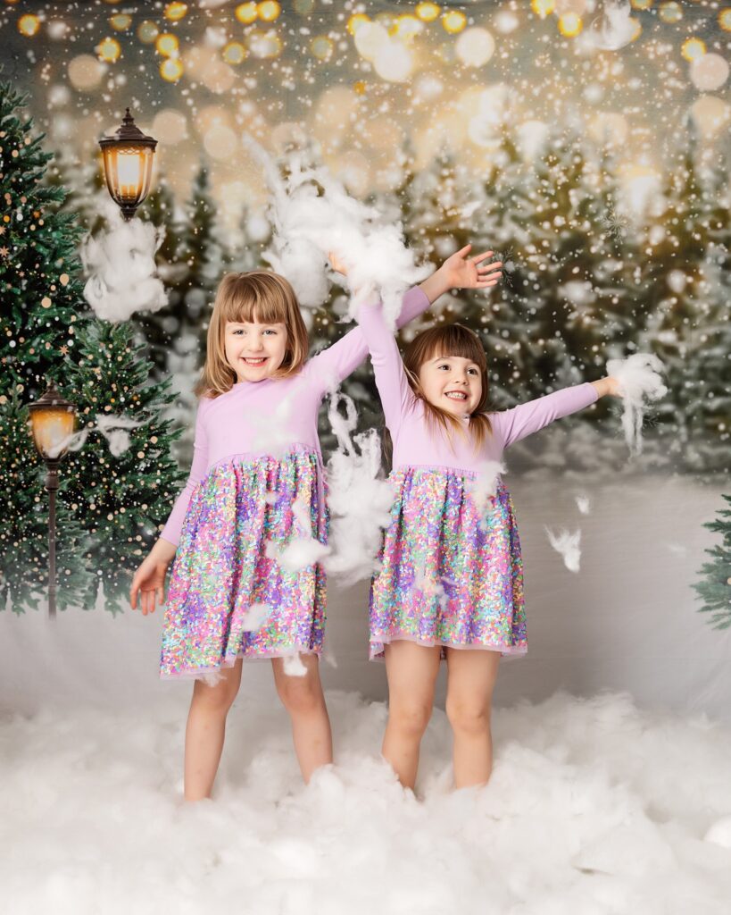 Two little girls wearing pink, lilac and green pretty dresses. Playing in the faux snow throwing it in the hair. Background scene, faux christmas woodland scene lit by street lamps.
