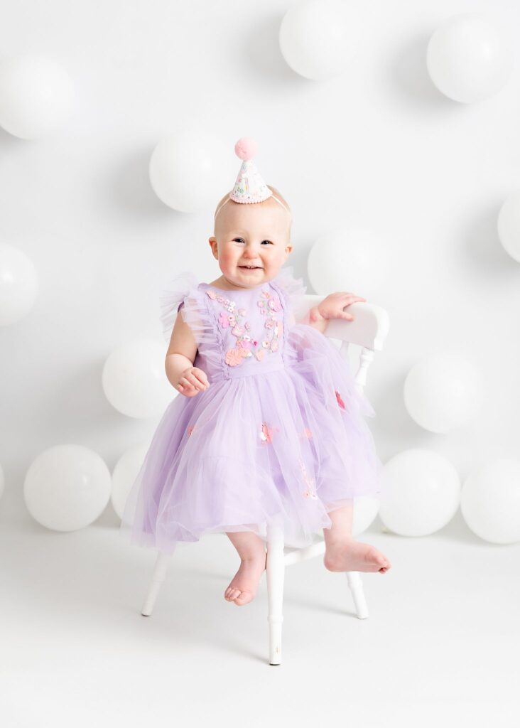 Sweet little girl in purple. First birthday photoshoot on a white set with white balloons. Pretty dress and number 1 cone hat. Taken at Meagan Sarah Photography studios for her cake smash session. Horsforth, Leeds.