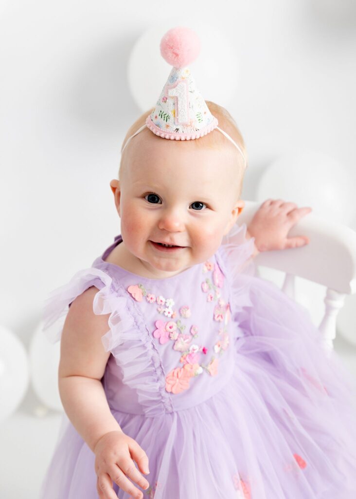 Sweet little girl in purple. First birthday photoshoot on a white set with white balloons. Pretty dress and number 1 cone hat. Taken at Meagan Sarah Photography studios for her cake smash session. Horsforth, Leeds. Sitting on a white chair.