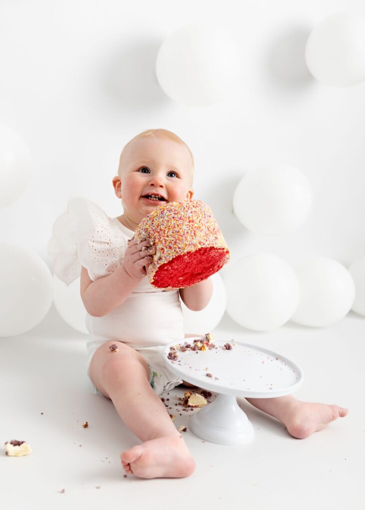 Pretty little baby girl. First birthday cake smash. Eating a rainbow cake from M&S food. Wearing a white frilly Zara kids vest. Photoshoot on a white set, white background, cake stand and white balloons. Happy baby. Taken at Meagan Sarah Photography studios Leeds. Professional photographer Leeds, Horsforth. West Yorkshire.