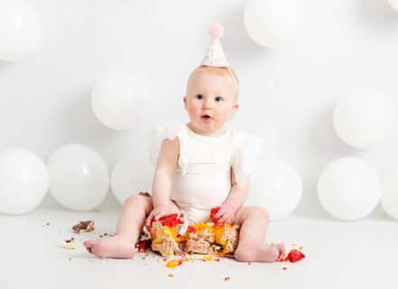 Pretty little baby girl. First birthday cake smash. Eating a rainbow cake from M&S food. Wearing a white frilly Zara kids vest. Photoshoot on a white set, white background, cake stand and white balloons. Happy baby. Taken at Meagan Sarah Photography studios Leeds. Professional photographer Leeds, Horsforth. West Yorkshire. Eating cake.
