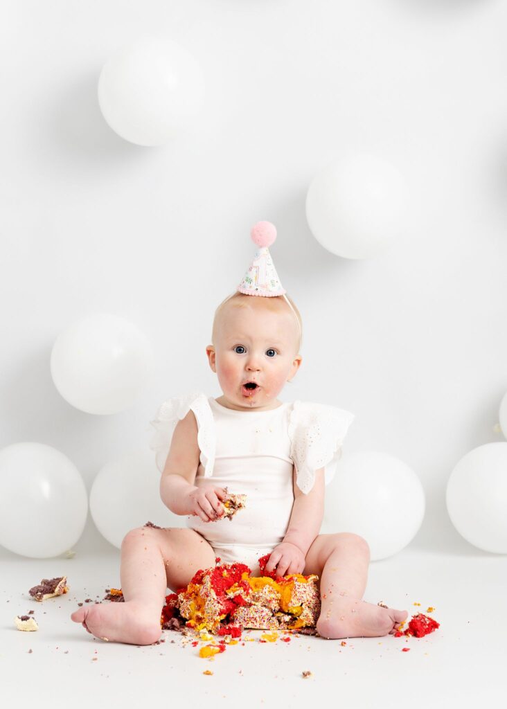 Pretty little baby girl. First birthday cake smash. Eating a rainbow cake from M&S food. Wearing a white frilly Zara kids vest. Photoshoot on a white set, white background, cake stand and white balloons. Happy baby. Taken at Meagan Sarah Photography studios Leeds. Professional photographer Leeds, Horsforth. West Yorkshire. Eating cake.