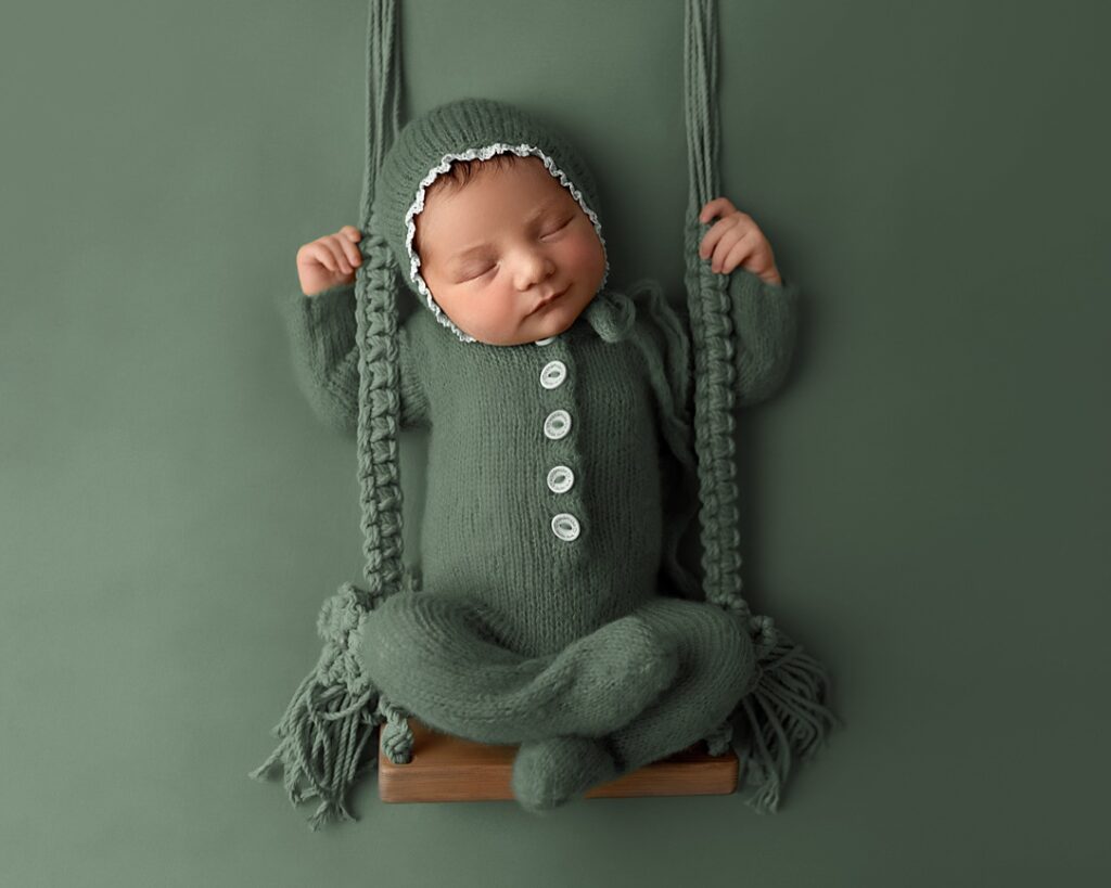 Baby photographer Leeds. Beautiful newborn photography Leeds. Photo of a newborn baby sitting on a wooden and macrame swing. Wearing a knitted button down onsie and knitted bonnet. All one colour of sage green.