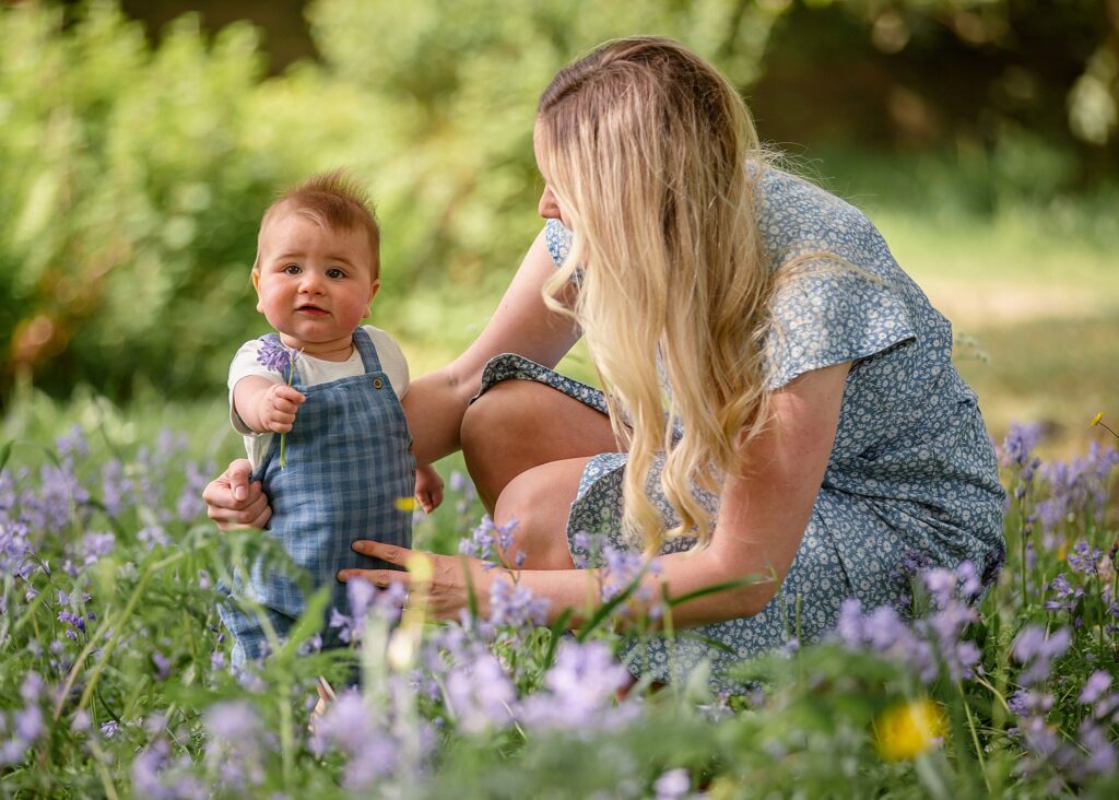 Part of our motherhood mummy and me photo sessions in the park. this is an image of a mother, with long blonde hair and a blue dress, sitting in the long grass with her young toddler watching him playing in the purple flowers amongst the long green grass. Image taken at Kirstall abbey, Leeds. Outdoor photoshoots. family portraits. 