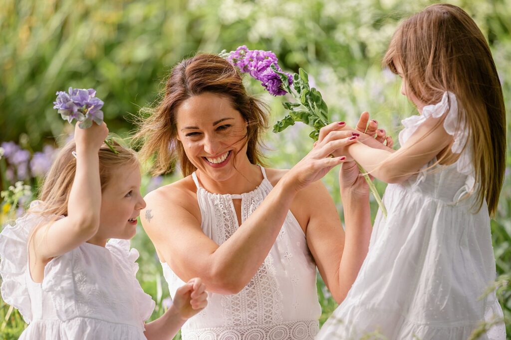 Part of our motherhood mummy and me photo sessions in the park. this is an image of a mother with her two young daughters in the long green grass. all in white dresses play fighting with purple flowers. Image taken at Kirstall abbey, Leeds. Outdoor photoshoots. family portraits. 