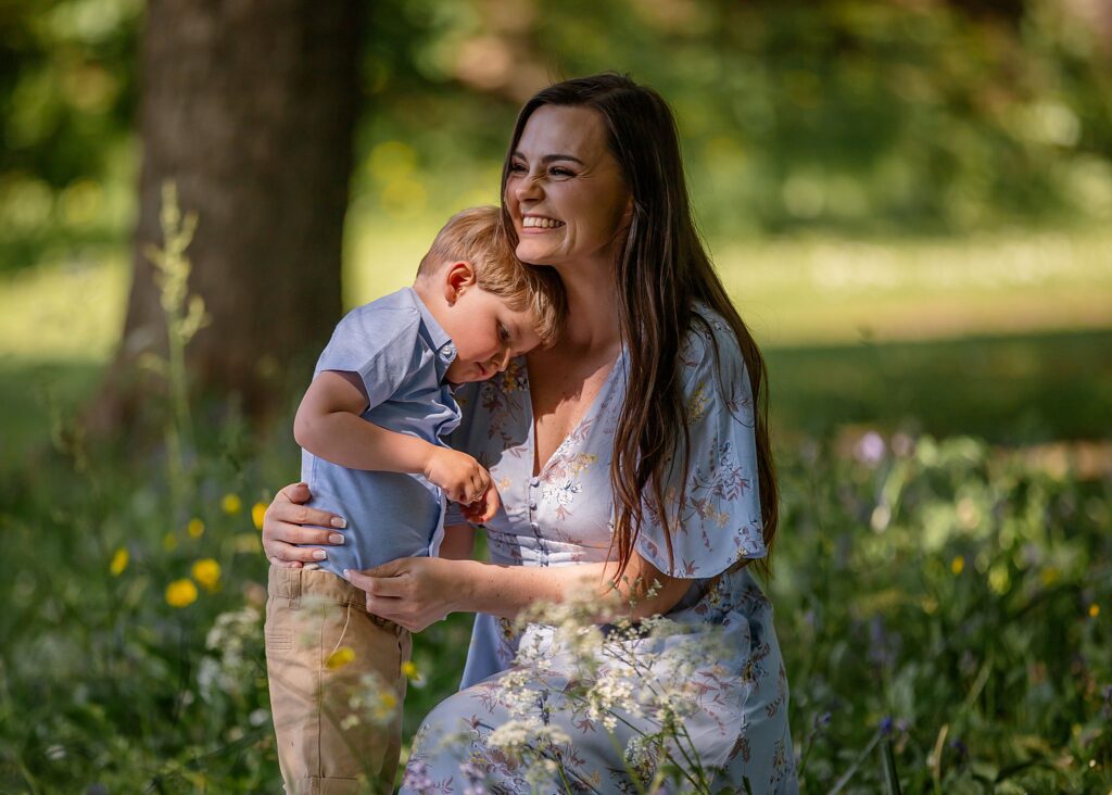 A beautiful woman embracing her son with a smile. Sitting in the shade of a tree with spots of lights on their face. sitting in the long grass with buttercups and cow parsley. Wearing blue. this portrait photoshoot is taken at Kirkstall Abbey, Leeds.
outdoor photoshoot.