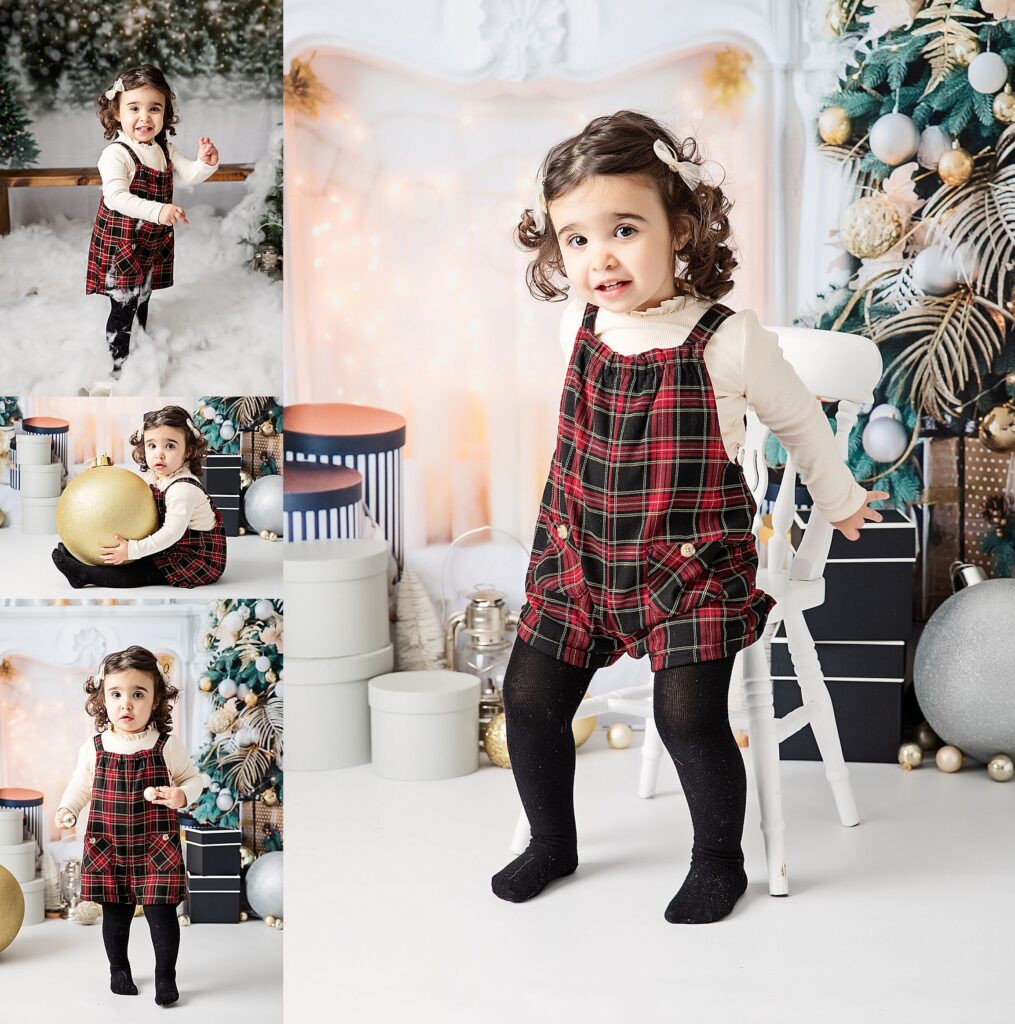 Pretty little girl with curly, shoulder length hair neat with a hair clip. Red and black flannel play suit with a white long sleeved top and black tights. Posing for her christmas photos taken against a faux christmas scene studio background. A glowing fireplace lit by fairy lights, surrounded by baubles, gifts and a Christmas tree. Holding large gold bauble. Having lots of fun. Taken at Leeds photography studio.