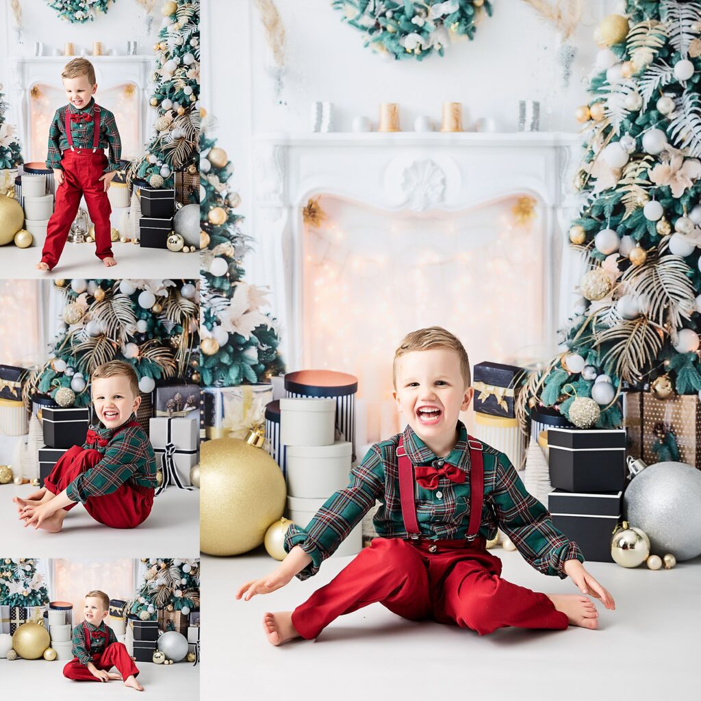 Little boy posing for his christmas photoshoot. Sitting against a faux christmas scene studio backdrop. Backdrop is of a fireplace lit with fairy lights surrounded by presents and baubles. A tree stands the the right. He is smiling and having fun whist sitting on the floor. Little boy wearing red trousers, red braces, red bowtie and a green flannel shirt.
