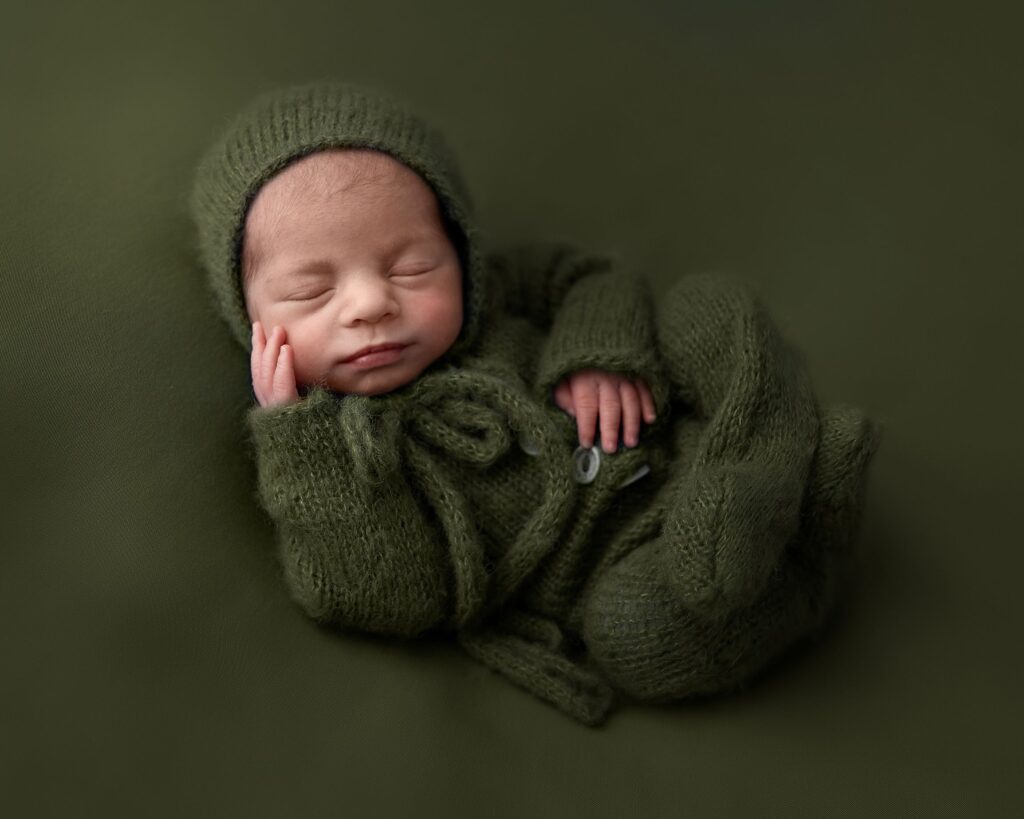 cute newborn baby boy asleep at his photoshoot. Laying on soft green fabric wearing a green knitted onesie and a bonnet tied in a bow. taken in Leeds photography studio by a professional photographer.